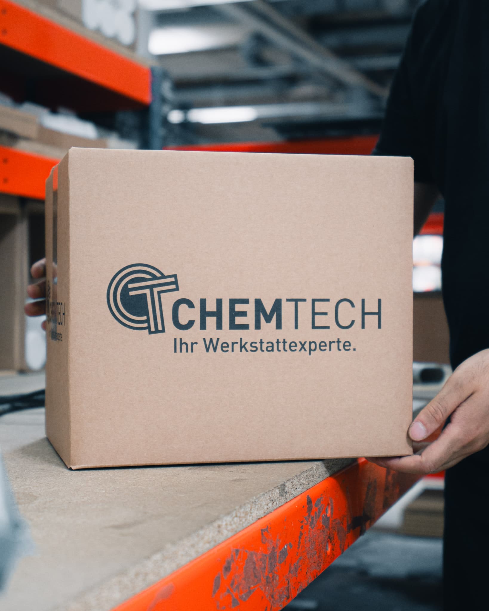 Zwei Hände reichen einen Karton mit ChemTech-Logo und dem Zusatz „Ihr Werkstattexperte“ über eine Regalkante im Lager