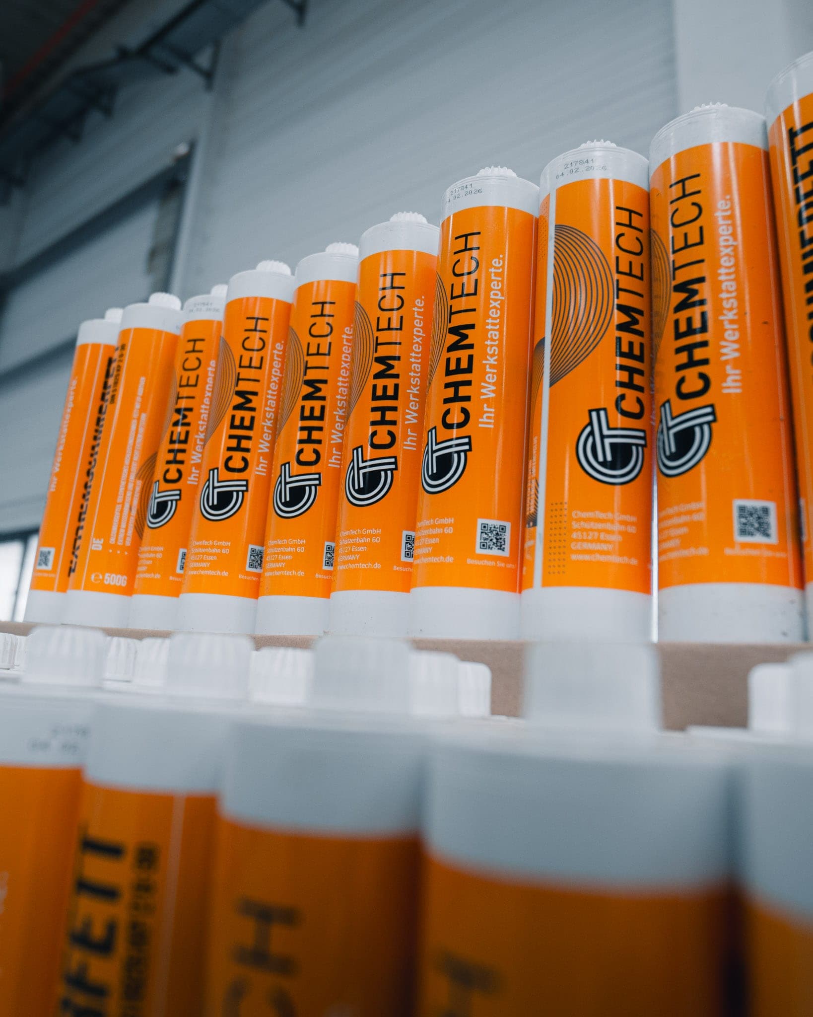 Reihen oranger ChemTech-Kartuschen mit Logo und dem Zusatz „Ihr Werkstattexperte“, dahinter weitere Gebinde in der Halle